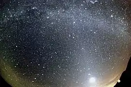 A meteor strikes the bottom left, while the Milky Way arcs overhead and a dawn-like light lines the lower horizon. The image was taken through a curved lens.