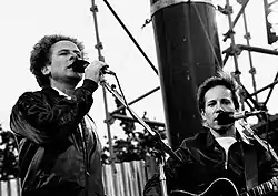 O cantaire y musico estausunidense Paul Simon, en una imachen d'os anyos 1980 chunto con Art Garfunkel en una actuación en os escenarios de Simon and Garfunkel.