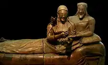 A sculpture of a woman and man reclining together on a couch, their upper bodies to the right and their legs to the left. There is a marked contrast between the high relief busts of their upper bodies and the very flattened lower bodies and legs. They have almond-shaped eyes and long braided hair, and are smiling widely. The man has a beard that is bobbed. The woman's hands are gesticulating in front of her as if she was holding something that is no longer there, or perhaps gesturing while speaking. The man has his right arm draped around the woman's shoulders in an intimate pose, and his right hand on her shoulder also appears to once have held some item. His left hand rests palm up in the crook of the woman's left elbow.