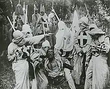 A black-and-white screenshot of several men in white robes and hoods surrounding an African-American man (a white actor using "blackface"). The men's robe have crosses on them and in the background other men are on top of horses.