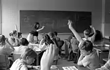 Students are seated at tables in a classroom. Some students have their hands raised and a teacher is pointing to one of these students.