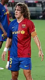 Curly haired man, looking tired, wearing a red and blue football shirt. In the background green grass is rarely visible.