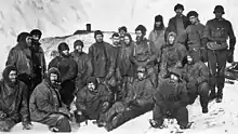 A group of men sitting closely packed together in heavy winter clothes and hats. Snow and ice surrounds them.