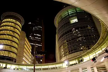 Night image of several tall skyscrapers taken from a street view, looking up. Several lights and traffic lights can be seen on the street, along with a round walkway above the street.