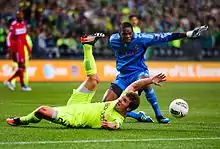 A player in a blue goalkeeper uniform stands with his hands extended over a player in a yellow uniform sliding on the ground. A ball is bouncing away from the players.