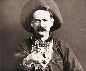 A black-and-white screenshot of a man facing the camera, while also pointing a revolver forward. He has a large mustache and is wearing a cowboy hat, a bandana around his neck, and a heavy jacket.