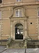 Un escalier de six marches conduit à une porte dont l'encadrement est orné par deux colonnes surmontée d'un fronton.