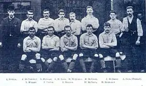 A black and white photograph of a group of men. The men in the front are sitting down, while the row behind they are standing