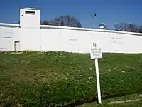 A tall white-painted wall with an overlapping round top and a square turret on the left-hand side. Behind it is a grey watchtower with an octagonal top and a searchlight on the roof. Further back are a group of white-painted half-timbered buildings and several trees.