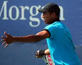 A man with blue T-shirt, black cap and tennis racquet in right hand