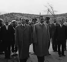 Two men standing side-by-side in the forefront, wearing overcoats. Behind them are several men in military uniform or suits and ties standing and saluting or making no gestures.