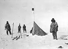 Four figures in heavy clothing stand near a pointed tent on which a small square flag is flying. The surrounding ground is ice-covered. Ski and ski poles are shown on the left.