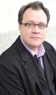 A bespectacled man in a black jacket, waistcoat, and tie, pink shirt, and jeans, sitting with his back to a marble-effect wall.