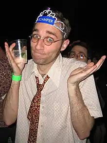 A young man wearing a loosened paisley tie and holding a plastic party cup shrugging at the camera.