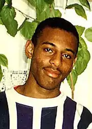 Photograph of a young Afro-Caribbean male, cropped to show his chest and head.  He has black hair, shaved very short, and a slight moustache.  He is wearing a navy and white vertically-striped crew neck shirt.  He is standing in front of a large indoor plant.