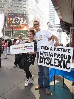 A girl holding a sign that says "LD
= equally intelligent / Cross out stigma" poses for a photo in Times Square with a man holding a sign that says "Take a picture with a proud Dyslexic".