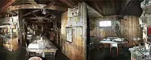 Dimly lit interior of a timber structure with a long table on which are papers and other unidentifiable objects. There are also bunks, sheles and packing cases. To the right, under a small window, is a smaller table with a chair.