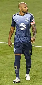 A shaven-headed man in a red football shirt with white sleeves, shorts and socks, applauding the crowd.