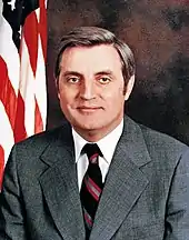 A man with dark hair, wearing a gray jacket, white shirt, and black striped tie