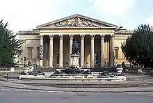 A Palladian style nineteenth century stone building with a large colonnaded porch. In front a large metal statue on a pedestal and fountains with decorations.