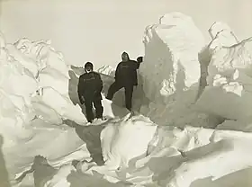 Two men in heavy clothing stand surrounded by mounds of ice which extend well above the height of their heads