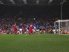 People in blue and red shirts on a field with a ball in the air. In the background is a stand that contains a lot of people.