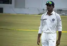 An Asian cricketer in cricket whites, wearing a dark blue baseball cap, with sunglasses on top. He is standing on his own on a cricket pitch.