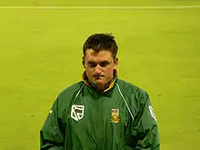 A white man with short dark hair in a green all-weather jacket. He is standing in front of a large expanse of grass.