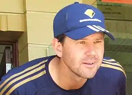 A white man with stubble, wearing a dark blue baseball cap with three white stripes on the peak and a yellow logo on the front. He is wearing a dark blue top with three yellow stripes down each arm from the shoulder and is leaning forward in front of a doorway.