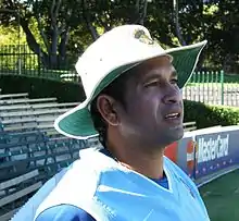 A man with dark skin in a light blue sleeveless pullover and dark blue t-shirt facing to the right. He is wearing a wide-brimmed white hat and is standing in front of some empty bleachers with trees further behind.