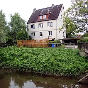 Situation 2013 am ehemaligen Mühlenstandort mit Wohngebäude aus den 1920er Jahren, an dessen Westseite ein altes Stadtwappen mit der Inschrift „1522“ eingelassen ist. Im Vordergrund der alte Okerverlauf vor Neubau des Ölper Wehrs in den 1970er Jahren.