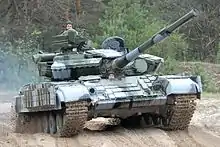 Vor einem Wald steht ein Panzer mit zwei Soldaten, die aus ihren Ausstiegsöffnungen blicken.