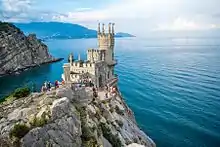 Vogelperspektive auf die Festung, die auf die Klippe gebaut wurde. Die Festung besteht aus hellem Stein und mehreren kleinen Türmen, die auf dem großen runden Turm und dem eckigen Gebäude verteilt sind. Viele Touristen haben sich auf der Plattform vor der Festung versammelt. Im Hintergrund sieht man das Meer und die Felsenküste.