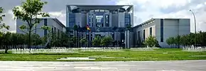 Frontale Farbfotografie vom Bundeskanzleramt mit Rasen und vielen kleinen Wasserfontänen. Vor dem Eingang stehen eine Europaflagge und eine Deutschlandflagge. Das Hauptgebäude besteht aus viel Glas und einigen Betonstützen und wird von zwei quaderförmigen Seitenflügeln flankiert.