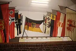 Links und rechts je 5 Flaggen an Stöcken, hinten an der Wand die aktuelle Dienstflagge der Deutschen Marine, vorne Tafeln mit Erläuterungen zu jeder Flagge