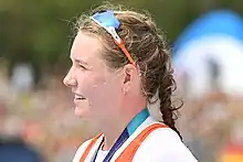 Tessa Dullemans schaut nach rechts. Sie trägt das orangene Nationaltrikot über einem weißen T-Shirt. Um den Hals hängt ein blaues Band, an dem die nicht sichtbare Silbermedaille befestigt ist. Ihre Sonnenbrille hat sie hochgeschoben.