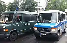 Frontale Farbfotografie von zwei Einsatzwagen der Polizei mit Fahrern.  Der linke Wagen hat eine dunkelgrüne Farbe und der rechte Wagen eine blau-silber gestreifte Farbe.