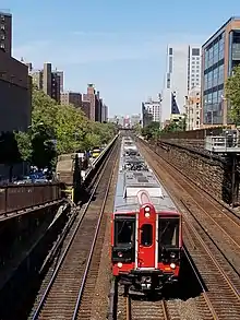 Großstadtszenerie: Rechts und links mehrstöckige Gebäude, in der Mitte eine viergleisige Eisenbahnstrecke, rechts mit gemauerter Stützwand, links mit Brüstung. Auf dem zweiten Gleis von links fährt ein rot-schwarz lackierter Triebwagenzug auf die Kameraposition zu, die sich auf einer Querstraße oberhalb der Gleise befindet und den Blick schräg aufs mit silbrigen Riffelblechen verkleidete Dach des Zuges fallen lässt.