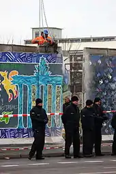 Frontale Farbfotografie von der Entfernung der Berliner Mauer mit schwarz gekleideten Polizisten und einem Absperrband im Vordergrund. In der Mitte ist ein Mauerstück nach hinten versetzt worden und ein Bauarbeiter befestigt eine Hebevorrichtung mit Ketten an dem linken Mauerstück an.