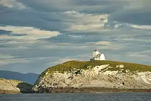 Foto einer grasbedeckten Insel mit einem weißen Leuchtturm darauf und bewölktem Himmel im Hintergrund