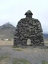 Das an einem bewölkten Tag aufgenommene Foto zeigt eine aus dunklen, teils flechtenbewachsenen Steinen aufgeschichtete Skulptur in der Form eines massiven Bogens mit einem pilzförmigen Auswuchs obendrauf.