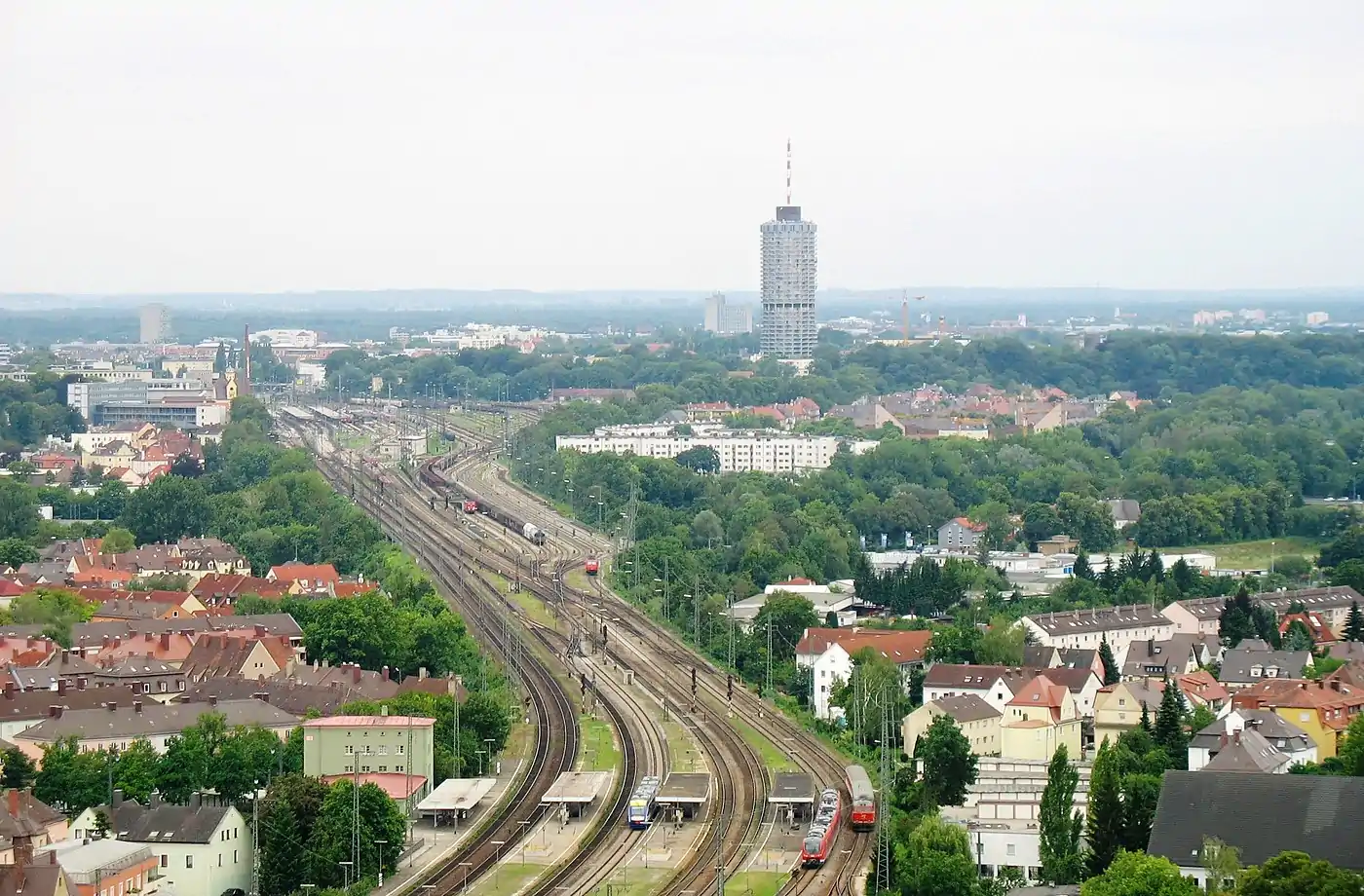 Links der Rand des Gleisfeldes am Augsburger Hauptbahnhof, viergeschossige optisch unterteilte weiße lange Gebäude im Baumbestand: Lessing- und Schubert Hof, dahinter das Thelottviertel.