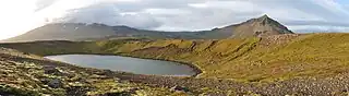 Das Panoramafoto zeigt einen sonnenbeschienenen, von Geröll und Moos bedeckten Krater, der etwa zur Hälfte von einem See ausgefüllt wird; im Hintergrund sind eine Gebirgskette und Wolken zu sehen.