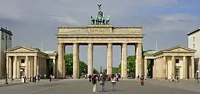 Frontale Farbfotografie vom Brandenburger Tor mit sechs Stützsäulen und einer Quadriga auf dem Dach. An beiden Seiten des Tors stehen zwei kleine klassizistische Gebäude mit Giebelfeldern. Auf dem Vorplatz laufen einige Menschen.