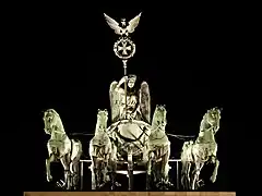 Nachtaufnahme einer Quadriga mit einem Stab in der Hand, das einen Ring mit Kreuz und darüber einen Adler trägt. Die Figur auf dem Streitwagen hat Flügel und trägt einen Lorbeerkranz sowie eine Toga. Im Vordergrund vier vorgespannte Pferde.