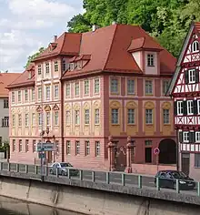 Seitliche Farbfotografie eines rosa, dreigeschossigen Gebäudes mit einem breiten und fünf kleinen Dachgiebeln. Die Fenster im ersten Stockwerk sind mit gelben Ornamenten versehen und die drei mittleren Fenster haben Giebelfelder. Rechts ist ein Eisentor von Steinpfeilern mit runden Vasenskulpturen gesäumt und daneben steht ein weißes Landhaus mit roten Balken. Die vordere Straße führt einen Fluss entlang.