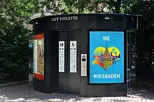 Zu sehen ist eine barrierefreie Toilettenanlage im öffentlichen Raum von Wiesbaden, die von Wall konzipiert und produziert wurde. Außen an der Toilettenanlage sind Plakatvitrinen zu sehen, die mit Plakaten bestückt sind.