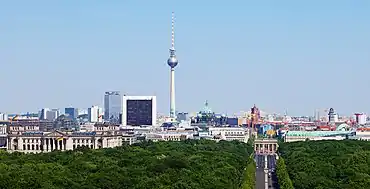 Farbfotografie von Berlin in der Vogelperspektive. Im Vordergrund stehen bewaldete Flächen vom Park Tiergarten und die Straße des 17. Juni führt zum Brandenburger Tor. Von links nach rechts sind das Reichstagsgebäude, der Fernsehturm, der Berliner Dom, das Rote Rathaus und das Brandenburger Tor zu sehen.