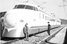 Schwarz-Weiß-Aufnahme vom zweiteiligen Zug A der JNR-Baureihe 1000 aus dem Jahr 1963. Der Triebzug steht auf freier Strecke und zwei Bahnangestellte, einer in Anzug und einer uniformiert, stehen vor dem vorne sichtbaren Endwagen A-021, während ein dritter Angestellter sich bei bei der Führerstandstür des anderen Endwagens befindet.