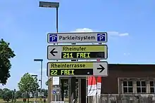 Blick auf drei längliche weiße Schilder. Ganz oben mit schwarzer Schrift „Parkleitsystem“, auf beiden Seiten ein blaues Rechteck mit einem weißen P unter einem weißen Dach. Das Schild darunter weist mit einem schwarzen Pfeil nach links, daneben steht auf gelbem Grund „Rheinufer“. Unter diesem schwarzen Schriftzug ist eine rechteckige schwarze Anzeige angeordnet. Sie zeigt mit einer blockigen hellgelben Schrift „211 FREI“ an. Das Schild darunter ist gleich aufgebaut, weist aber nach rechts. Die Schrift lautet oben „Rheinterrasse“, unten „22 FREI“. Hinter den Schildern erkennt man den Himmel, einige Bäume und Gebäude.